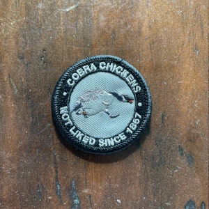May include: A black and white embroidered patch with the text "Cobra Chickens Not Liked Since 1997" and a white goose with an orange beak.