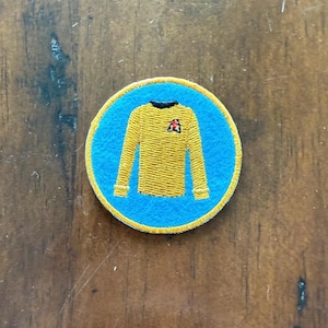 May include: A blue and yellow embroidered patch featuring a yellow Star Trek uniform with a red delta symbol on the chest.