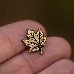 May include: A small, detailed gold-colored maple leaf pin. The pin features intricate veining and a small, rounded stem. The design is set against a blurred background, highlighting the pin's delicate features.