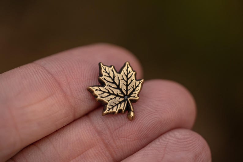 May include: A small, detailed gold-colored maple leaf pin. The pin features intricate veining and a small, rounded stem. The design is set against a blurred background, highlighting the pin's delicate features.