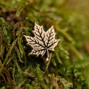 May include: A bronze-colored maple leaf pin with detailed veining, set against a backdrop of green moss. The pin's design is intricate, showcasing the natural form of the leaf. The overall aesthetic is rustic and nature-inspired.