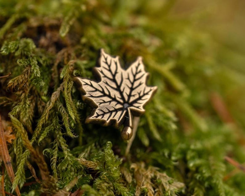 May include: A bronze-colored maple leaf pin with detailed veining, set against a backdrop of green moss. The pin's design is intricate, showcasing the natural form of the leaf. The overall aesthetic is rustic and nature-inspired.