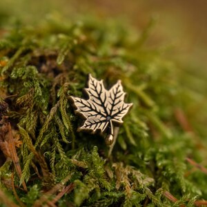 May include: A detailed, antique-style maple leaf pin with intricate vein detailing. The pin is made of a gold-toned metal and is set against a backdrop of lush green moss. The pin's design captures the essence of autumn.