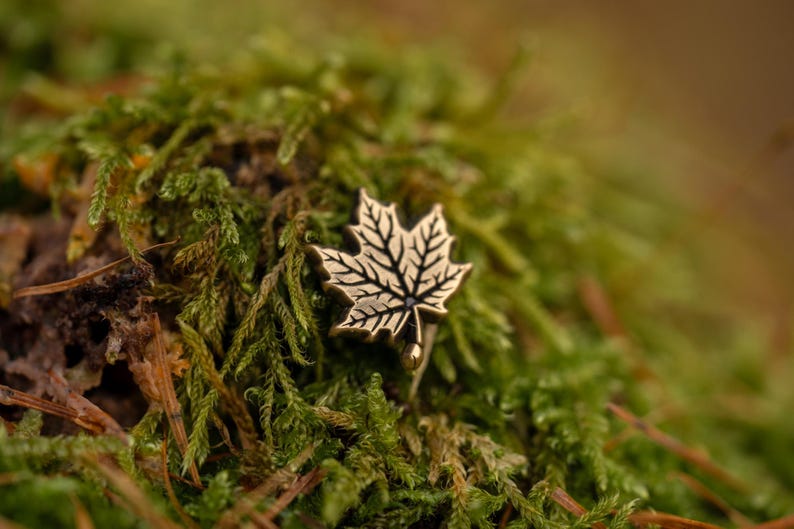 May include: A detailed, antique-style maple leaf pin with intricate vein detailing. The pin is made of a gold-toned metal and is set against a backdrop of lush green moss. The pin's design captures the essence of autumn.