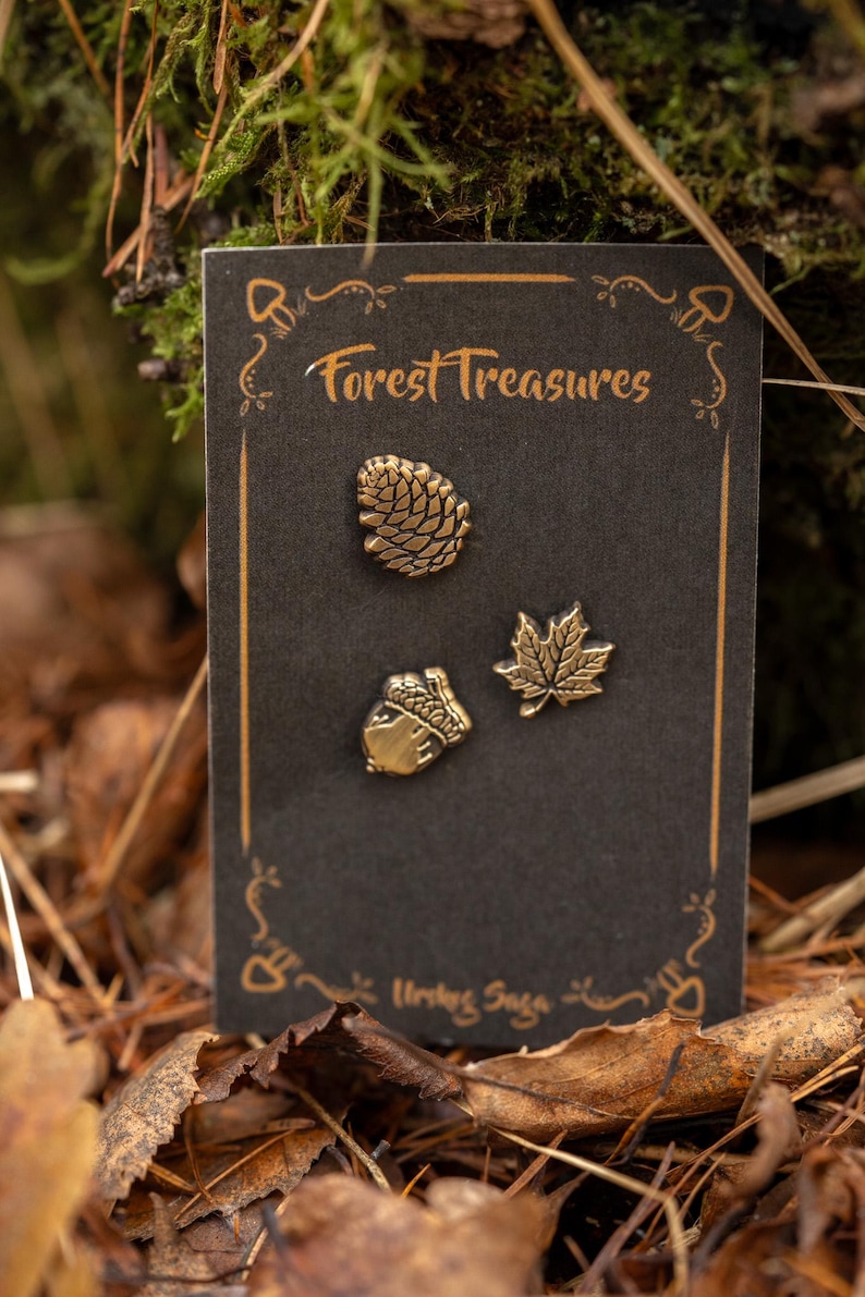 May include: A black card with the text "Forest Treasures" and three gold-colored pins: a pinecone, an acorn, and a maple leaf. The card is set against a backdrop of leaves and moss.