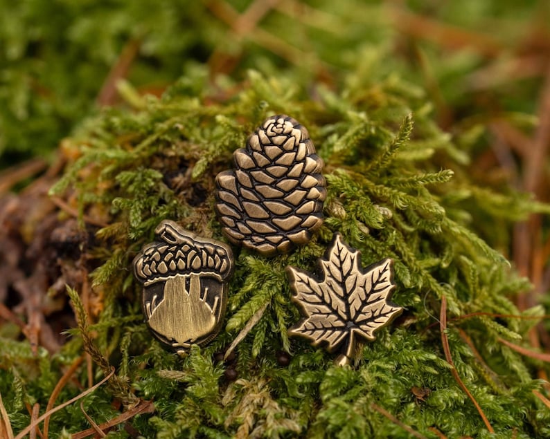 May include: Three bronze-colored pins: a pine cone, an acorn, and a maple leaf. The pins are set against a backdrop of green moss and brown twigs, creating a natural, earthy aesthetic. The detailed designs of the pins are highlighted by the contrasting textures.