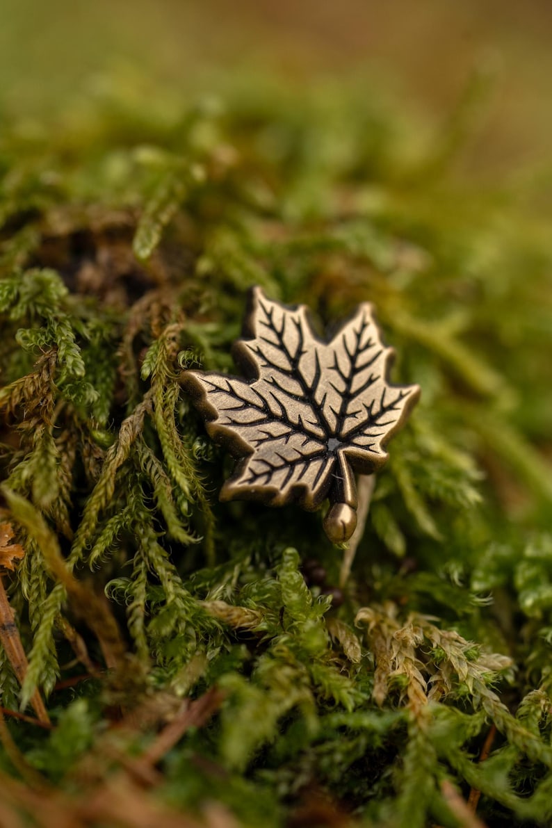 May include: A bronze-colored maple leaf ring set against a backdrop of green moss. The ring features detailed veining on the leaf, with a smooth, rounded base. The natural setting highlights the ring's organic design.