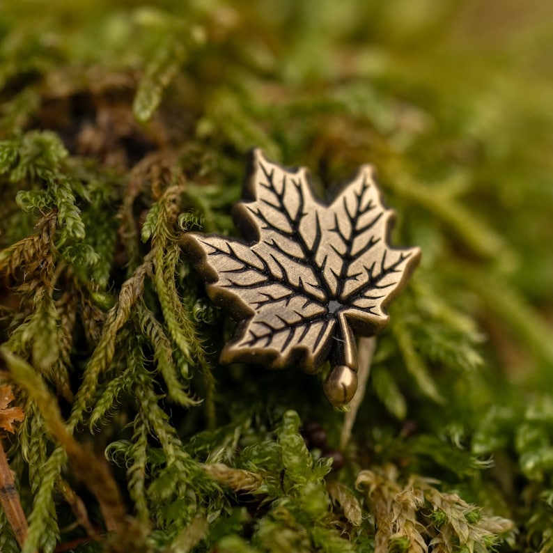 May include: A bronze-colored maple leaf pin with detailed veining, set against a backdrop of green and brown moss. The pin has a small, dark center and a rounded base. The image is a close-up, highlighting the texture and detail of the pin and moss.