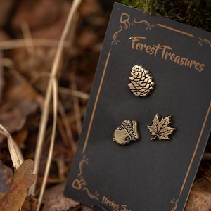 May include: A black card with three bronze-colored pins: a pine cone, an acorn, and a maple leaf. The card reads "Forest Treasures" in a decorative font. The background is blurred with leaves and foliage.