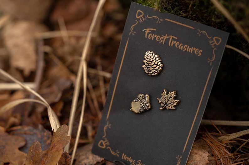 May include: A black card with three bronze-colored pins: a pine cone, an acorn, and a maple leaf. The card reads "Forest Treasures" in a decorative font. The background is blurred with leaves and foliage.