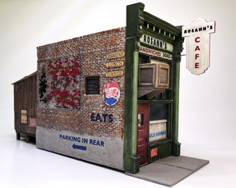 May include: A miniature model of a vintage cafe, featuring a brick facade with weathered signage for "Rosann's Cafe," "Sandwiches Dogs," and "Old Drinks." A Pepsi logo and "EATS" are visible. The model includes a "Parking in Rear" sign.
