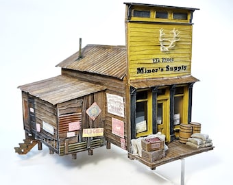 May include: A model of a wooden building with a sign that reads "Elk River Miner's Supply". The building has a yellow facade with black trim and a brown roof. There are several signs on the building, including one that says "Old Gold".