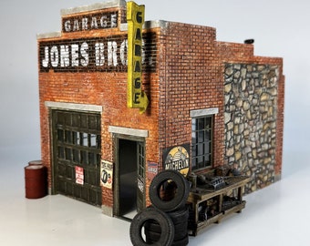 May include: A model of a brick garage with a sign that reads "Garage Jones Bros." and a yellow sign that reads "Garage". The garage has a wooden door and a window. There is a stone wall on the right side of the garage. There are tires stacked in front of the garage.