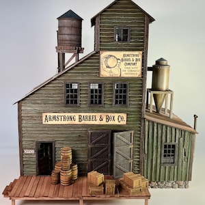 Pode incluir: Um modelo detalhado do edifício Armstrong Barrel & Box Co., com revestimento verde desgastado, barris de madeira e caixas numa plataforma de madeira. A placa diz "Armstrong Barrel & Box Co." e "Made with the Strongest Materials."
