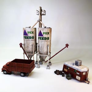 May include: A model train set with two white silos labeled "Apex Feeds", a brown truck, a red and white trailer labeled "Rent", and a pile of brown barrels.