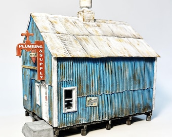 May include: A miniature model of a plumbing supply shop. The building has blue corrugated metal siding with rust-colored streaks. A red sign reads "PLUMBING & SUPPLY". The roof is made of weathered metal. A concrete step leads to the white door.