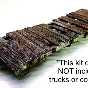 May include: A weathered model train flatcar with a wooden deck, featuring a detailed, aged appearance. The deck is made of brown planks with visible wear and tear. A black metal wheel and other mechanical details are visible. The text on the image says "This kit does NOT include trucks or couplers."