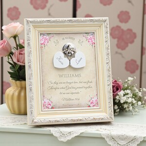 Könnte beinhalten: Ein gerahmtes Hochzeitsandenken mit einem cremefarbenen, verzierten Rahmen. Das Kunstwerk zeigt zwei herzförmige Schlösser mit eingravierten Namen, die miteinander verbunden sind. Der Rahmen enthält das Datum 18.10.2025, die Namen Kevin und Laura und ein Zitat aus Matthäus 19,6.