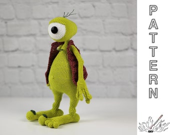 May include: A green crocheted monster with one large eye and a maroon scarf. The monster is standing on a white surface.
