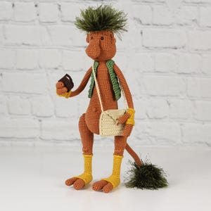 May include: A crocheted amigurumi toy of a brown creature with green hair, wearing a green vest and a beige bag. The toy is holding a small brown object in its hand.