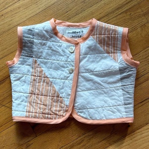 May include: A white and peach quilted vest with a button closure. The vest has a peach trim and a label that reads "Nugget's Knits".