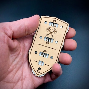 May include: A wooden game tracker with a shield shape, featuring engraved text and symbols. The tracker displays numerical values for CP, VP, AP, and RD, along with a crossed hammer emblem. The design is in light brown with white and gray accents.