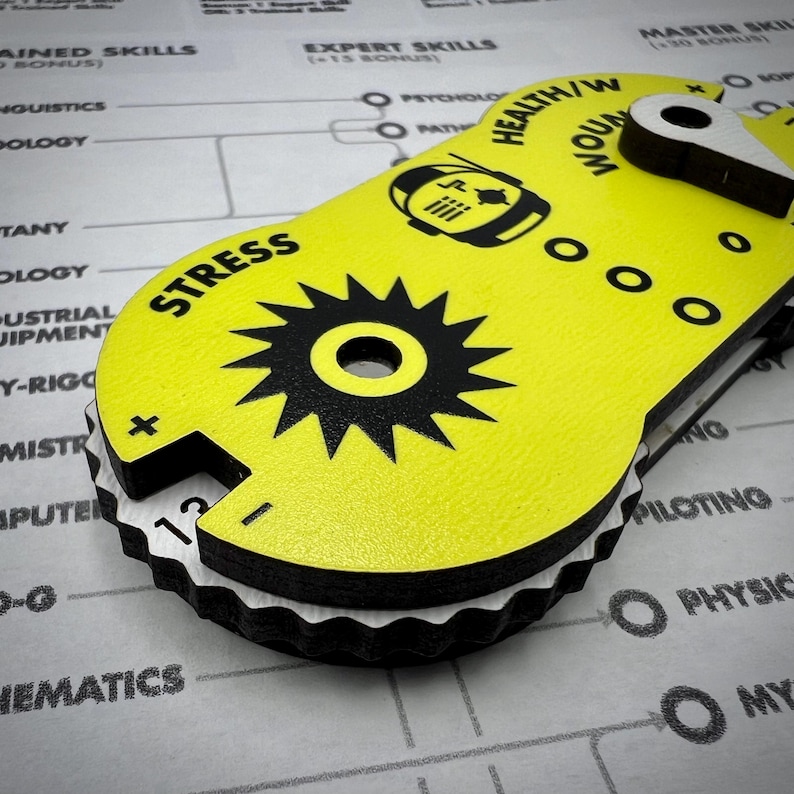 Puede incluir: Un rastreador de juego amarillo de forma ovalada con texto y gr&aacute;ficos impresos en negro. Cuenta con un dial central en forma de engranaje etiquetado como STRESS, una secci&oacute;n HEALTH/WOUND con un puntero blanco giratorio e indicadores.