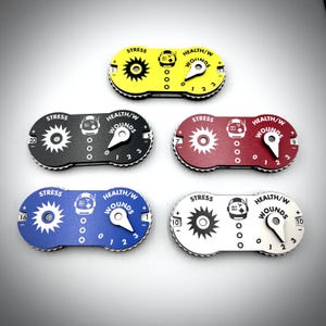Puede incluir: Cinco rastreadores de juego ovalados en amarillo, negro, rojo, azul y blanco. Cada uno presenta un dial de estr&eacute;s en forma de engranaje, un dial de salud y heridas con puntero, y un icono de casco de astronauta con el texto STRESS y HEALTH/W WOUNDS.
