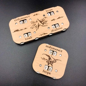 May include: Two wooden game pieces with engraved images and numbers. The first piece has a mountain scene with the words "Endurance 40", "Load 10", "Hope 19", and "Shadow 04". The second piece has a dragon image with the words "Endurance 39" and "Hope 12".