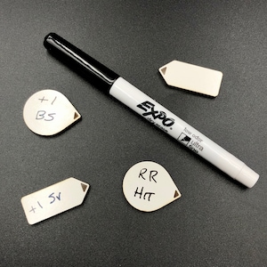 May include: A white dry erase marker with a black cap and the words "Expo" and "low odor ultra fine" printed on the side. The marker is lying on a black surface next to four white labels with handwritten text. The labels read "+1 BS", "RR HT", "+1 5V", and a blank label.