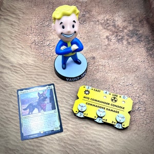 May include: A yellow and black plastic game piece with the text "MTG Commander Console" and "Commander Damage". The piece has a dial with numbers 0, 1, 2, 3, 4, 5, 6, and 7. The piece is next to a card with a dog on it and a small figurine of a cartoon character with yellow hair and a blue shirt.