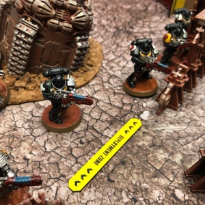 May include: A yellow sign with black text that reads "Deployment Zone" with arrows pointing to the right. The sign is on a brown, cracked surface. There are several miniature figures of space marines in black and silver armor with red and blue accents.