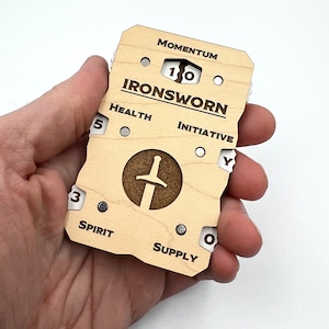 May include: A wooden character tracker for role-playing games. It features a sword icon, the words "IRONSworn", and dials for tracking Momentum, Health, Initiative, Spirit, and Supply.