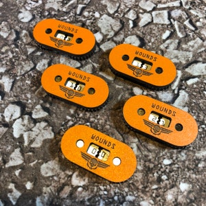 May include: Five orange and brown wooden counters with the word "WOUNDS" and a skull graphic. Each counter has a number from 0 to 3 displayed on it.