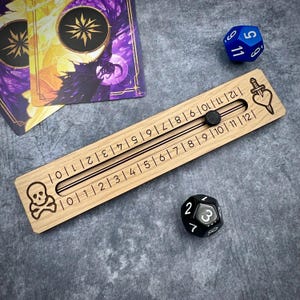 May include: A wooden game score tracker with a sliding bead, numbered from 0 to 12 on each side. A skull and crossbones is etched on one end, and a dagger through a heart on the other. Two dice and two game cards are also present.