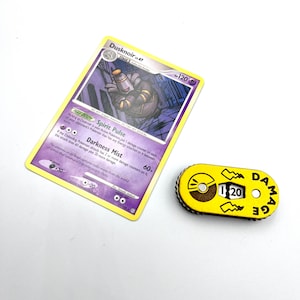 May include: A yellow Pokemon damage counter with a black and white Pokeball graphic and the number 120 printed on it. The counter is labelled "DAMAGE".