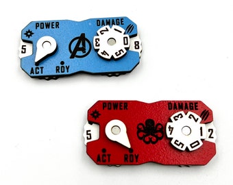 May include: Two red and blue plastic character tokens with dials for a board game. The blue token has a white dial with numbers 2, 4, 0, and 8. The red token has a white dial with numbers 7, 5, 3, 2, and 12. Both tokens have the words "POWER" and "DAMAGE" printed on them.