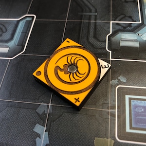 May include: A yellow square game piece with a black scorpion design and the letters "I", "E", "+", and "-" on the sides.