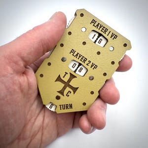 May include: A tan game board with the text "PLAYER 1 VP" and "PLAYER 2 VP" above two number wheels. The wheels display the numbers 16 and 08. The board also has a cross symbol and the word "TURN".
