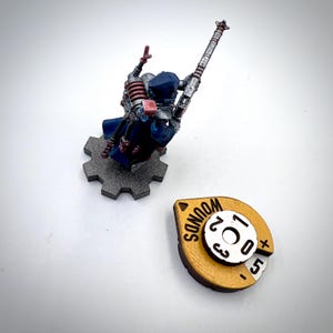 May include: A miniature painted figure of a robot warrior holding a weapon, standing on a grey gear-shaped base. A yellow and white wooden wound tracker with the text "Wounds" and numbers 1-5.
