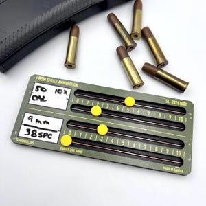 May include: An olive green Schooner Labs Prism Series Ammunition counter with yellow sliding markers, white labels reading 50 10x and 9mm 38SPC, and the text Danger Live Ammo. Five brass-cased cartridges are positioned above the device.
