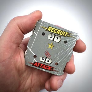 May include: A square grey game counter featuring the text RECRUIT above a 5 and 1, and ATTACK above a 0 and 2. The device includes a yellow star graphic, red claw marks, and white diagonal lines on a textured surface with mechanical dial windows.