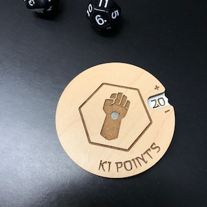 May include: A wooden K1 POINTS game token featuring a fist graphic within a hexagon. The token has a sliding dial displaying the number 20. Two black dice are visible in the background.