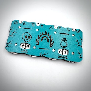 May include: A teal wooden game tracker with a skull, a ghost, and a potion symbol. It has rotating dials for "LIFE" and "MANA" with numbers. The tracker has a tree branch design along the edges and various symbols and numbers.