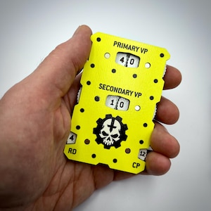 May include: A bright yellow rectangular game score tracker with black dots and a skull gear logo. It features windows displaying numbers for PRIMARY VP, SECONDARY VP, RD, and CP. The device has mechanical dials to adjust the numerical values shown.