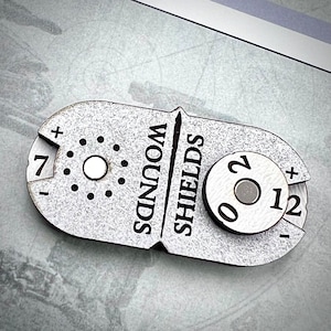 May include: A silver-coloured, two-sided tracker with a circular dial on each side. One side is labelled "Wounds" and has a dial with numbers 1 through 7. The other side is labelled "Shields" and has a dial with numbers 0 through 2.