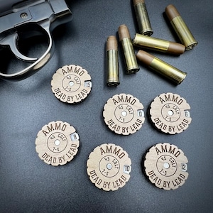 May include: Six wooden round tokens with the words "AMMO 45 COLT 7-5 DEAD BY LEAD" and a number from 0 to 12. The tokens are on a dark brown surface.
