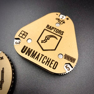 May include: A tan-coloured game token featuring the word "RAPTORS" above a shield emblem and the word "UNMATCHED" below. The token has a distinctive shape with cutouts and markings, including numbers and symbols.