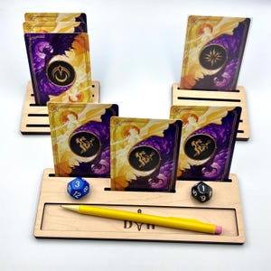May include: Wooden card holders with game cards featuring a purple and gold design. A yellow pen, a blue 12-sided die, and a black 6-sided die are in the foreground. The cards have a circular emblem and a sun-like background.