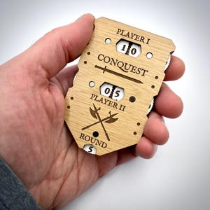 May include: A wooden game score tracker with the word "CONQUEST" and the text "PLAYER I" and "PLAYER II". The tracker displays the scores 10 and 05, and the round number 5. The design includes crossed axes and a sword.